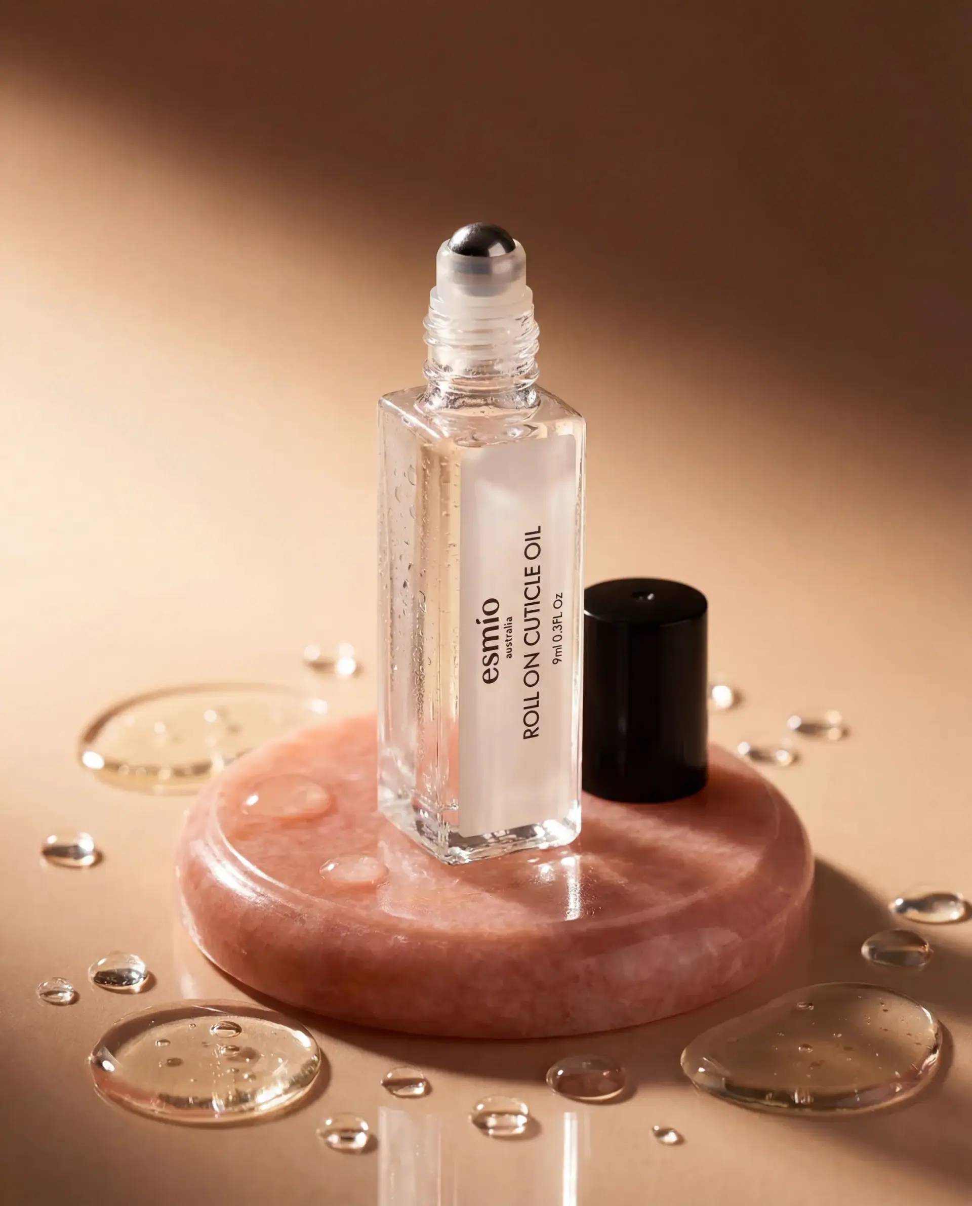 Ezmio cuticle-oil roll-on on a blush-pink stone plinth, scattered with water droplets